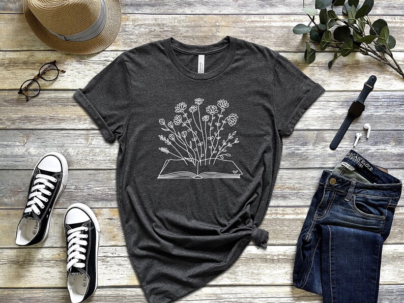 K&ouml;nnte beinhalten: Ein dunkelgraues T-Shirt mit einer wei&szlig;en Umrandung eines Buches, aus dem Blumen wachsen.