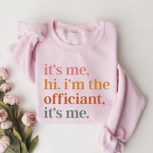 May include: Pink sweatshirt with the text "it's me, hi. i'm the officiant, it's me." printed in orange, red, and green.