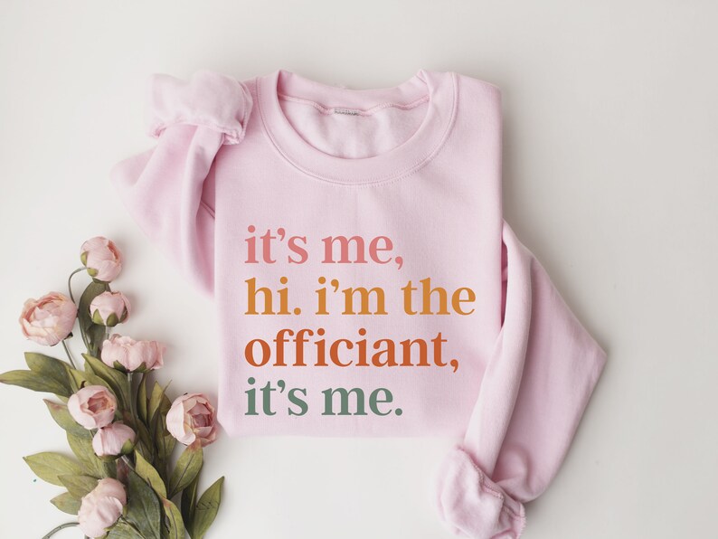 May include: Pink sweatshirt with the text "it's me, hi. i'm the officiant, it's me." printed in orange, red, and green.