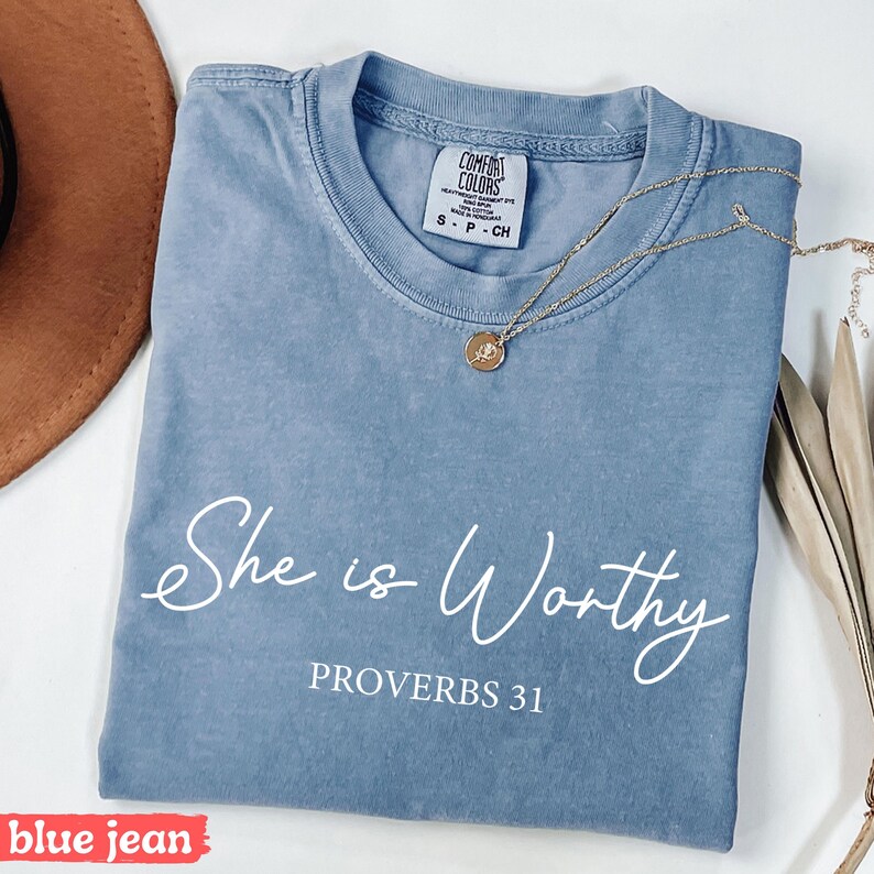 May include: Blue jean Comfort Colors t-shirt with the words "She is Worthy" and "Proverbs 31" in white script. A gold necklace with a pendant rests on the shirt. The shirt is folded.