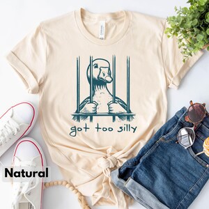 May include: A beige t-shirt with a graphic of a goose in a cage. The text "got too silly" is printed below the image.