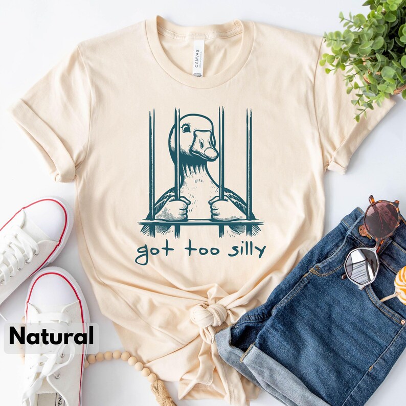 May include: A beige t-shirt with a graphic of a goose in a cage. The text "got too silly" is printed below the image.