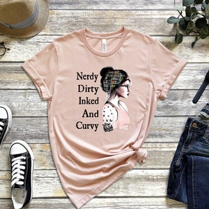 Op de afbeelding: Een roze T-shirt met een afbeelding van een vrouw met een boekenkast als haar, die een bril draagt en een tatoeage op haar arm heeft. De tekst op het shirt luidt "Nerdy Dirty Inked And Curvy".