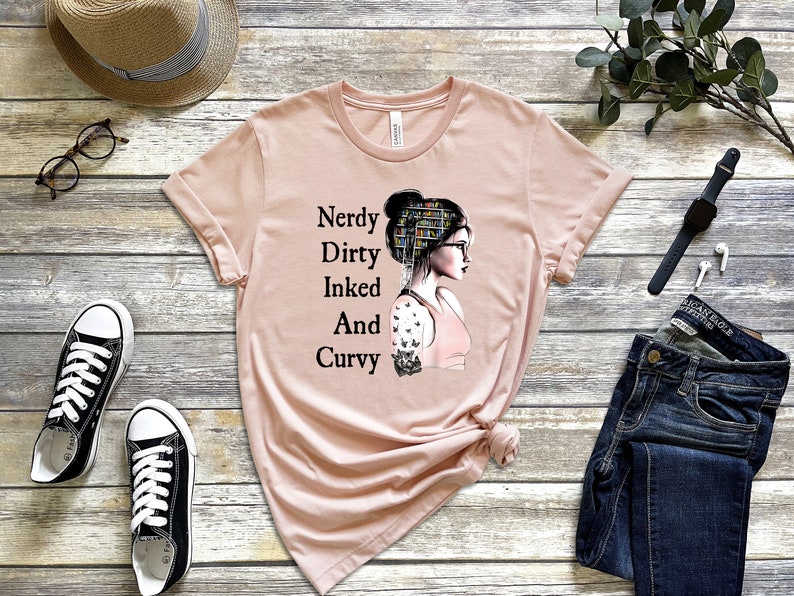 Op de afbeelding: Een roze T-shirt met een afbeelding van een vrouw met een boekenkast als haar, die een bril draagt en een tatoeage op haar arm heeft. De tekst op het shirt luidt "Nerdy Dirty Inked And Curvy".