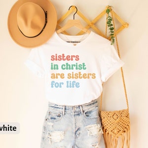 May include: White t-shirt with the text "sisters in christ are sisters for life" in rainbow colors. The shirt is on a hanger and is being displayed on a white wall.