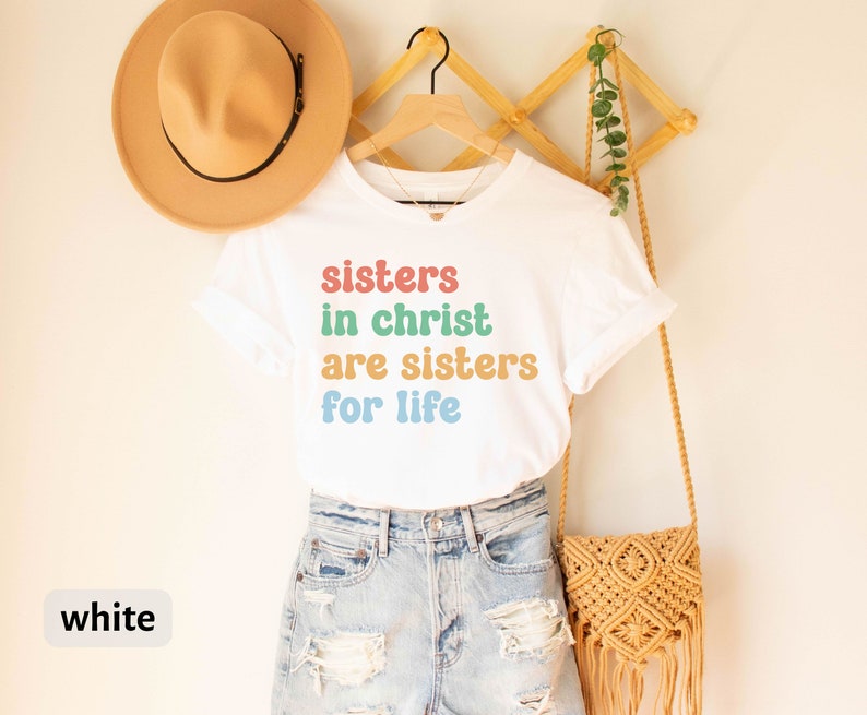 May include: White t-shirt with the text "sisters in christ are sisters for life" in rainbow colors. The shirt is on a hanger and is being displayed on a white wall.