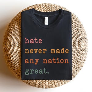 Op de afbeelding: Zwart T-shirt met de tekst "hate never made any nation great." gedrukt in oranje, roze en groen.