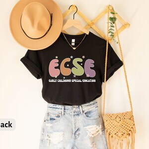 K&ouml;nnte beinhalten: Schwarzes T-Shirt mit dem Text "ECSE" in bunten Buchstaben, steht f&uuml;r "Early Childhood Special Education". Das Shirt h&auml;ngt an einem Holzgestell mit einem Strohhut und einer kleinen gewebten Tasche.