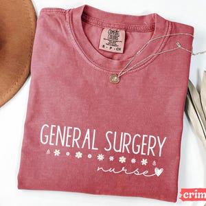Może przedstawiać: Czerwony t-shirt z napisem "GENERAL SURGERY nurse" w białym kolorze. Koszulka ma małe białe serce i akcenty w postaci stokrotek.