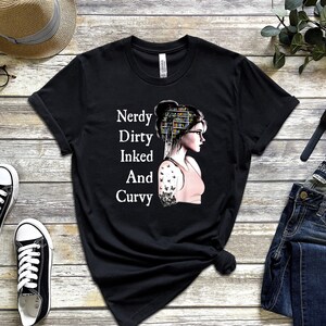 Op de afbeelding: Zwart T-shirt met een witte afbeelding van een vrouw met boeken op haar hoofd en een tatoeage op haar arm. De tekst op het shirt luidt "Nerdy Dirty Inked And Curvy".
