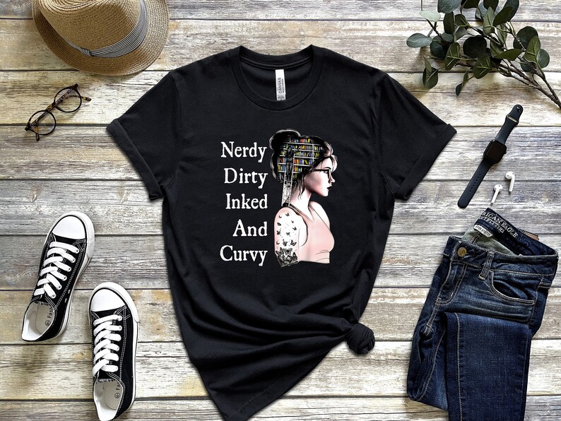 Op de afbeelding: Zwart T-shirt met een witte afbeelding van een vrouw met boeken op haar hoofd en een tatoeage op haar arm. De tekst op het shirt luidt "Nerdy Dirty Inked And Curvy".
