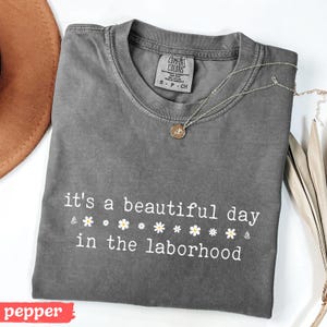 May include: A gray Comfort Colors t-shirt with the text "it's a beautiful day in the laborhood" in white. The shirt is folded and has a small tag and a gold necklace with a pendant. A brown hat is visible in the background.