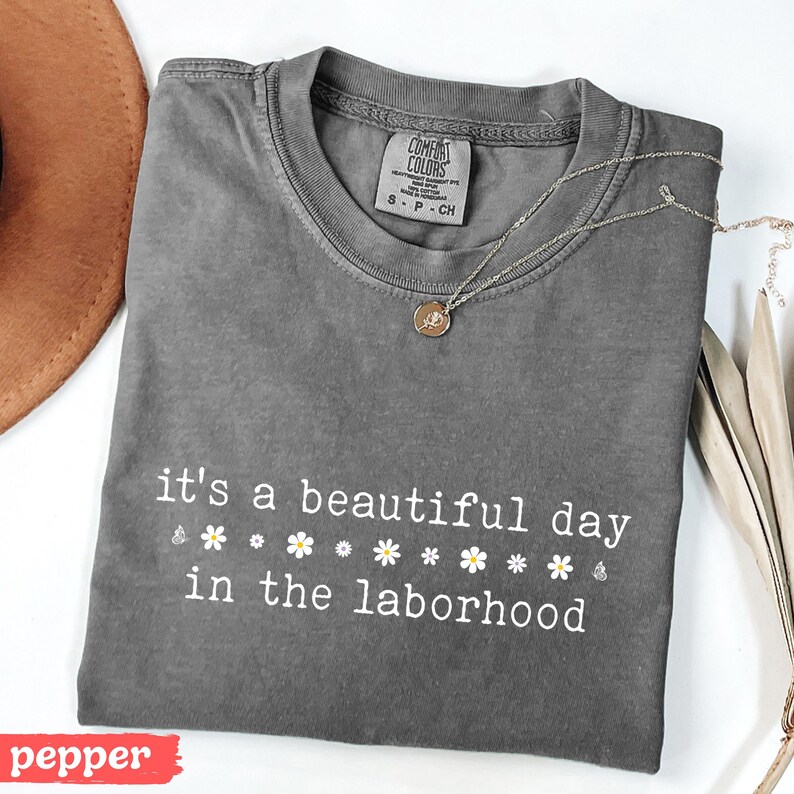 May include: A gray Comfort Colors t-shirt with the text "it's a beautiful day in the laborhood" in white. The shirt is folded and has a small tag and a gold necklace with a pendant. A brown hat is visible in the background.