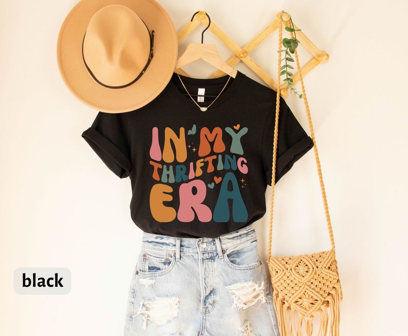 May include: Black V-neck t-shirt with the text "IN MY THRIFTING ERA" in colorful retro font. The shirt is displayed with a tan hat, a gold necklace, and a woven beige crossbody bag. The shirt is paired with light blue denim shorts.
