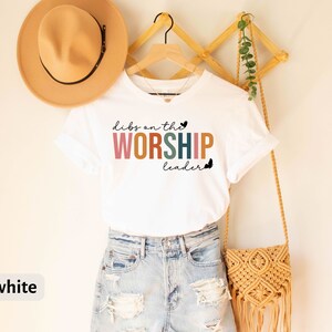 Puede incluir: Camiseta blanca con el texto "Dibs on the Worship Leader" en letras de colores. La camiseta est&aacute; en una percha y tiene un peque&ntilde;o coraz&oacute;n al lado del texto.