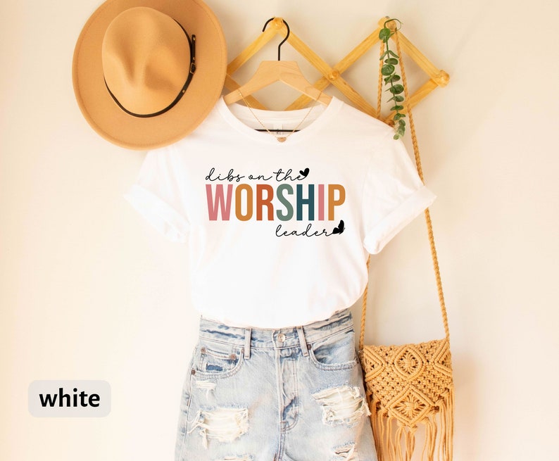 Puede incluir: Camiseta blanca con el texto "Dibs on the Worship Leader" en letras de colores. La camiseta est&aacute; en una percha y tiene un peque&ntilde;o coraz&oacute;n al lado del texto.