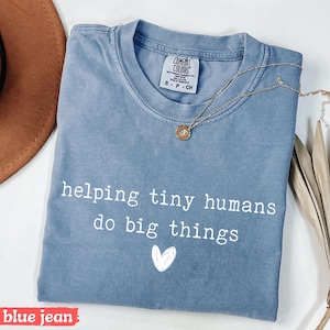 May include: Blue jean colored t-shirt with the text "helping tiny humans do big things" in white, along with a small white heart graphic. A gold necklace with a pendant is draped across the shirt.