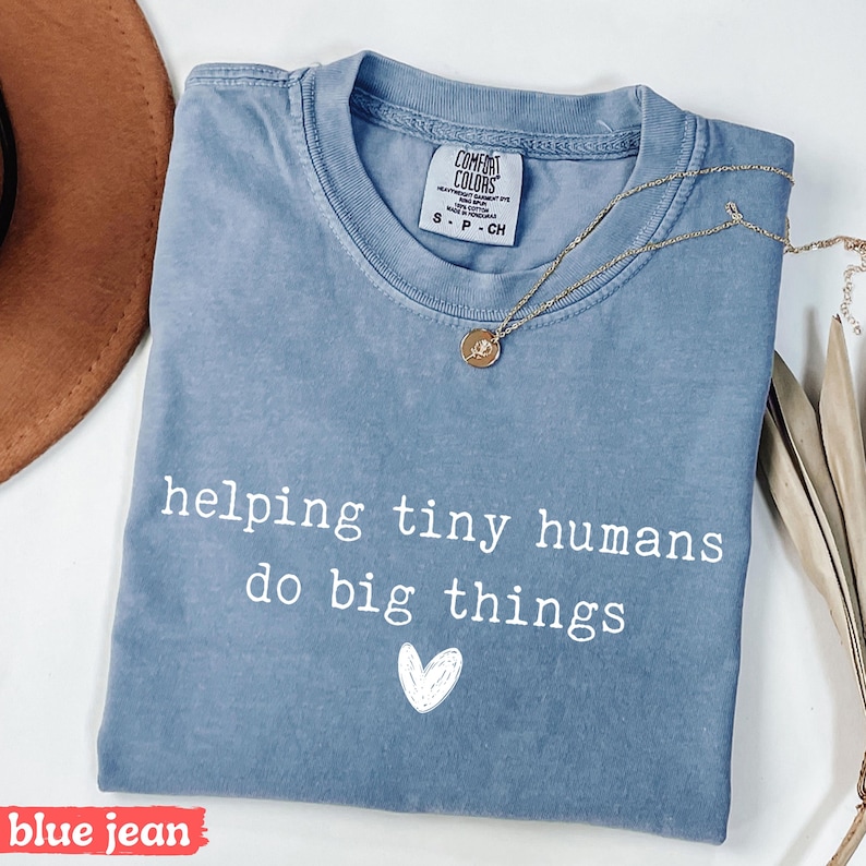 May include: Blue jean colored t-shirt with the text "helping tiny humans do big things" in white, along with a small white heart graphic. A gold necklace with a pendant is draped across the shirt.