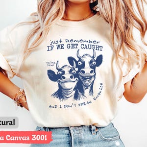 Może przedstawiać: Kremowy t-shirt z zabawną grafiką dw&oacute;ch uśmiechniętych kr&oacute;w w kolorze granatowym. Tekst brzmi "Just Remember If We Get Caught" i "And I Don't Speak English". Wykonany z materiału Bella Canvas 3001.