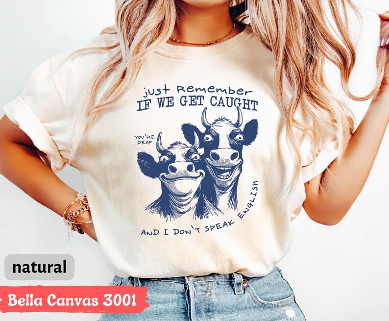 Może przedstawiać: Kremowy t-shirt z zabawną grafiką dw&oacute;ch uśmiechniętych kr&oacute;w w kolorze granatowym. Tekst brzmi "Just Remember If We Get Caught" i "And I Don't Speak English". Wykonany z materiału Bella Canvas 3001.