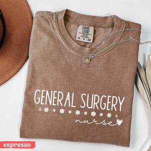 Może przedstawiać: Koszulka w kolorze espresso z napisem "GENERAL SURGERY nurse" w kolorze białym, z małym sercem i rzędem białych stokrotek.