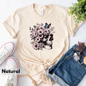 May include: A beige t-shirt with a graphic of a skull with pink flowers and a blue butterfly on top. The skull is facing to the right. The t-shirt is laid out on a surface with a pair of white sneakers, a pair of blue denim shorts, and a wooden bead necklace.