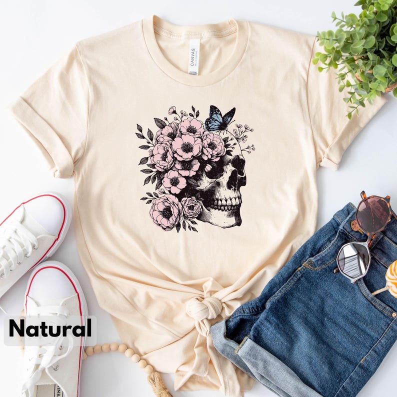 May include: A beige t-shirt with a graphic of a skull with pink flowers and a blue butterfly on top. The skull is facing to the right. The t-shirt is laid out on a surface with a pair of white sneakers, a pair of blue denim shorts, and a wooden bead necklace.