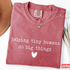 May include: Crimson-colored t-shirt with the text "helping tiny humans do big things" and a white heart graphic. A silver necklace with a pendant rests on the shirt. The shirt has a crew neck and a tag that says "Comfort Colors".