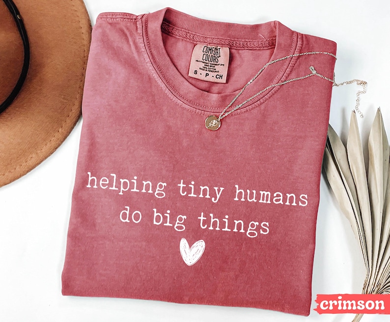 May include: Crimson-colored t-shirt with the text "helping tiny humans do big things" and a white heart graphic. A silver necklace with a pendant rests on the shirt. The shirt has a crew neck and a tag that says "Comfort Colors".
