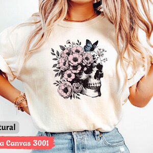May include: A white t-shirt with a graphic of a skull with pink flowers and a blue butterfly. The text "Bella Canvas 3001" and "natural" are printed on the bottom of the shirt.