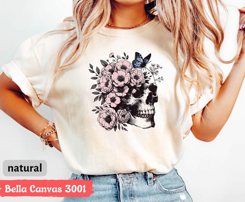 May include: A white t-shirt with a graphic of a skull with pink flowers and a blue butterfly. The text "Bella Canvas 3001" and "natural" are printed on the bottom of the shirt.