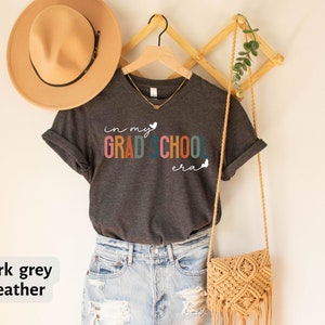 May include: Dark grey heather t-shirt with the text "in my grad school era" in rainbow colors with a heart and butterflies. The shirt is on a hanger with a straw bag and a hat.
