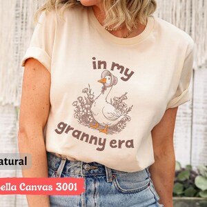 May include: A natural-colored Bella Canvas 3001 t-shirt with the phrase "in my granny era" printed on the front. The graphic features a cartoon goose wearing a bonnet and surrounded by flowers. The shirt has rolled-up sleeves.
