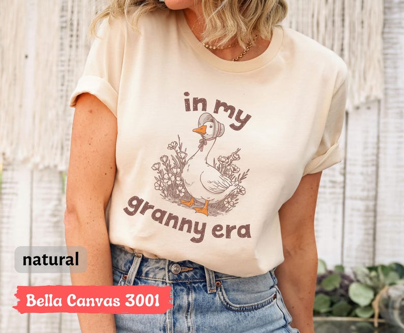 May include: A natural-colored Bella Canvas 3001 t-shirt with the phrase "in my granny era" printed on the front. The graphic features a cartoon goose wearing a bonnet and surrounded by flowers. The shirt has rolled-up sleeves.