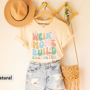 May include: A light beige t-shirt with the text "Weird Moms Build Character" in colourful, retro-style lettering. The shirt is hanging on a wooden hanger and has a natural colour.