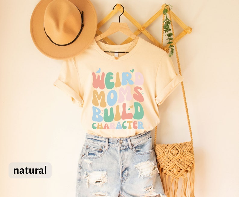 May include: A light beige t-shirt with the text "Weird Moms Build Character" in colourful, retro-style lettering. The shirt is hanging on a wooden hanger and has a natural colour.