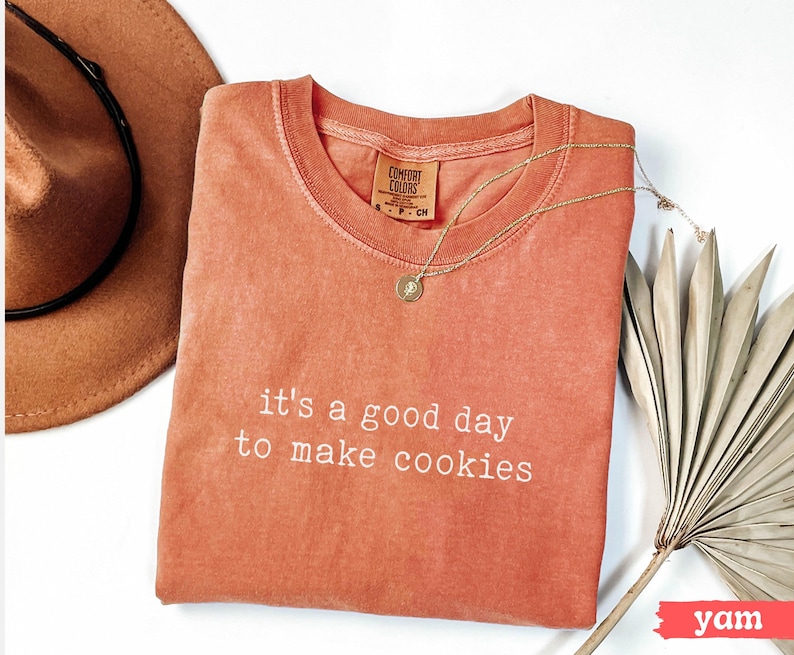May include: A coral coloured t-shirt with the text "it's a good day to make cookies" in white. The shirt is lying flat on a white surface with a brown hat and a dried palm leaf.