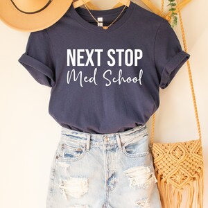May include: A navy blue t-shirt with the text "NEXT STOP Med School" in white lettering. The shirt is hanging on a wooden hanger.