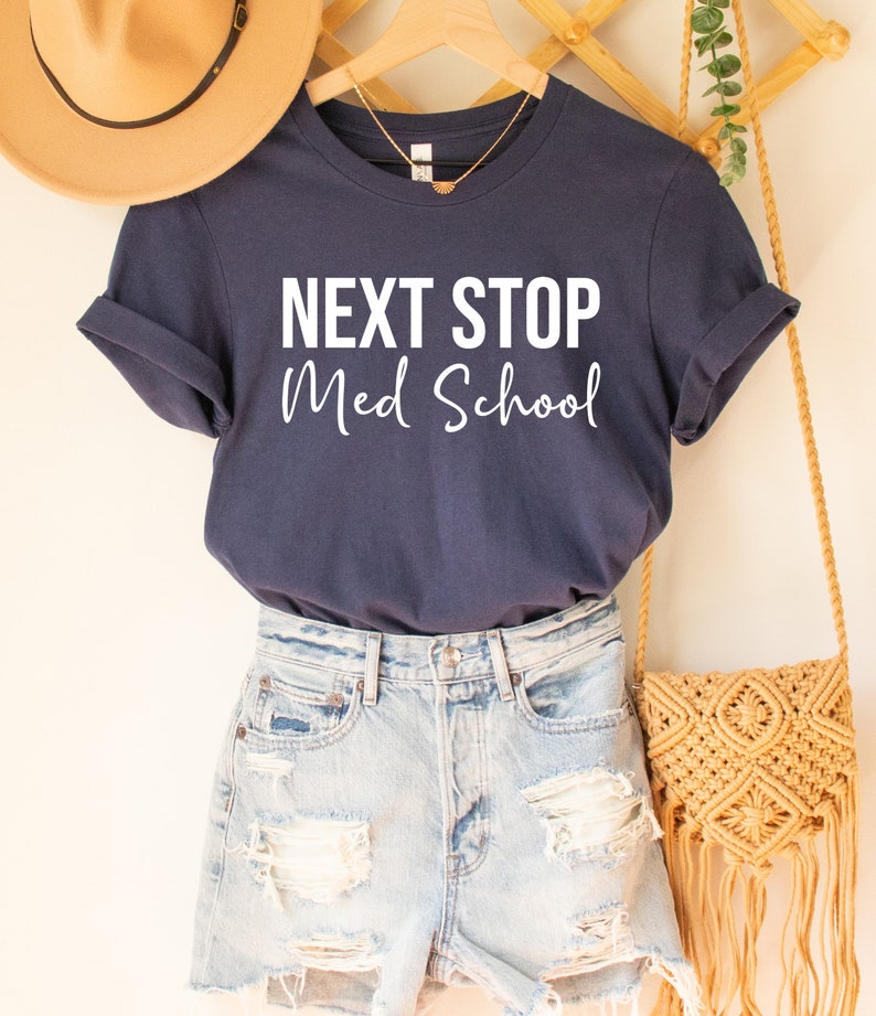 May include: A navy blue t-shirt with the text "NEXT STOP Med School" in white lettering. The shirt is hanging on a wooden hanger.