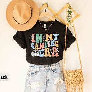 May include: Black t-shirt with the text "In My Camping Era" in colorful retro letters. The shirt features a small camper van graphic and is worn with denim shorts.