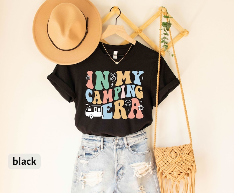 May include: Black t-shirt with the text "In My Camping Era" in colorful retro letters. The shirt features a small camper van graphic and is worn with denim shorts.