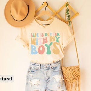 K&ouml;nnte beinhalten: Ein hellbeiges T-Shirt mit dem Text "Life is better with my boys" in bunten, Retro-Lettern. Das Shirt h&auml;ngt an einem Holzb&uuml;gel und ist mit einem Strohhut und einer geflochtenen Tasche gestylt.