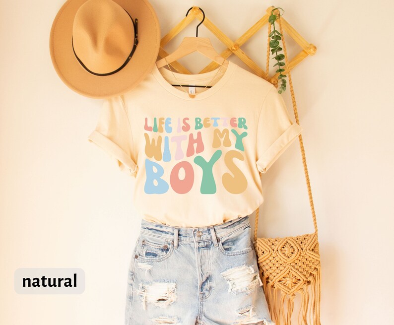 K&ouml;nnte beinhalten: Ein hellbeiges T-Shirt mit dem Text "Life is better with my boys" in bunten, Retro-Lettern. Das Shirt h&auml;ngt an einem Holzb&uuml;gel und ist mit einem Strohhut und einer geflochtenen Tasche gestylt.