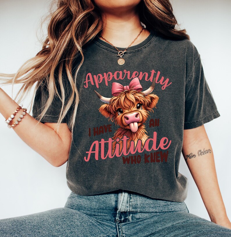 May include: A dark gray t-shirt with a distressed look featuring a cartoon Highland cow with a pink bow. The text "Apparently I have an Attitude Who Knew" is printed on the shirt in pink.