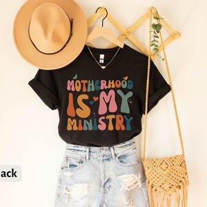 May include: Black t-shirt with the text "Motherhood is my ministry" in colorful retro lettering. The shirt is hanging on a wooden rack with a straw bag and a hat.