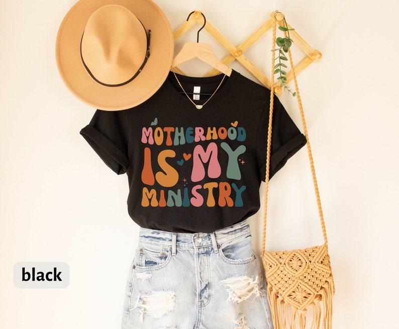 May include: Black t-shirt with the text "Motherhood is my ministry" in colorful retro lettering. The shirt is hanging on a wooden rack with a straw bag and a hat.