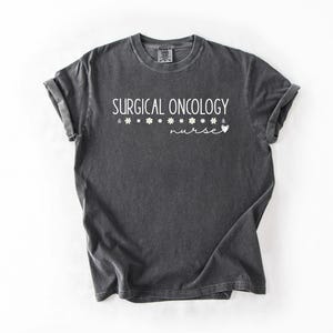 May include: A dark gray t-shirt with the text "SURGICAL ONCOLOGY nurse" printed in white. The text is surrounded by small white flower symbols.