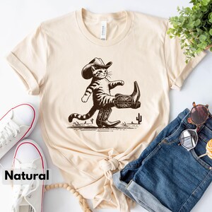 May include: Cream-colored t-shirt featuring a cartoon cat wearing a cowboy hat and boots, with a cactus graphic. The word "Natural" is printed at the bottom. The shirt is styled with blue jeans and white sneakers.