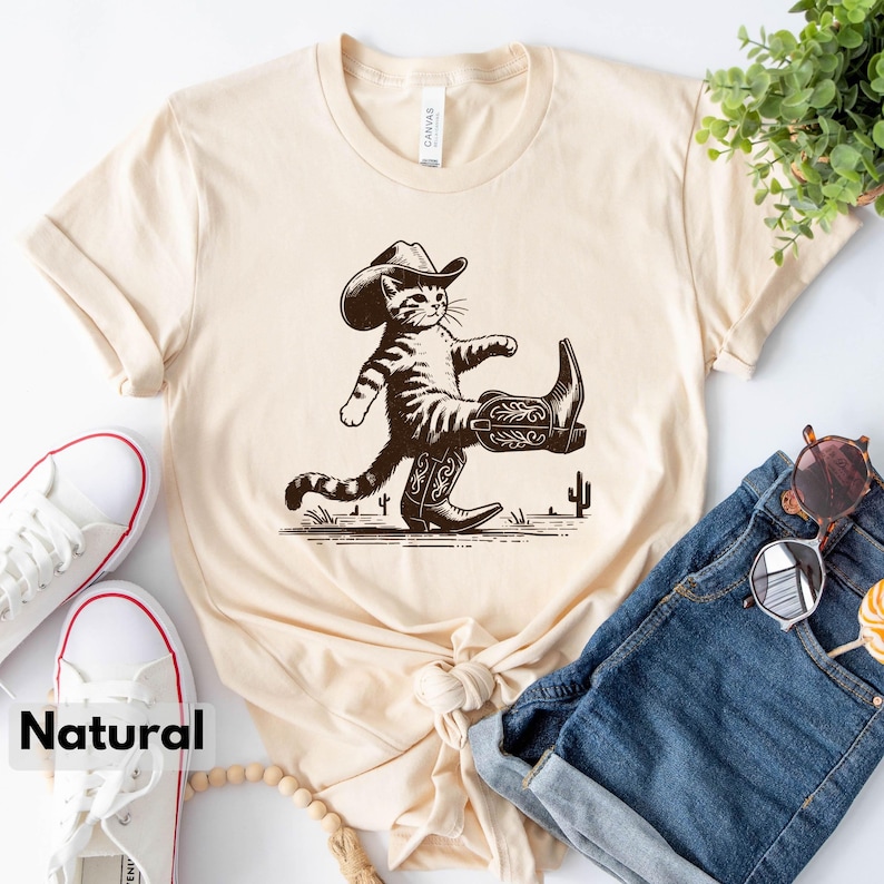May include: Cream-colored t-shirt featuring a cartoon cat wearing a cowboy hat and boots, with a cactus graphic. The word "Natural" is printed at the bottom. The shirt is styled with blue jeans and white sneakers.