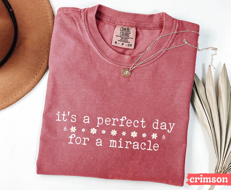 May include: A crimson red t-shirt with the text "it's a perfect day for a miracle" in white lettering. The shirt has a distressed look and features small white flowers around the text.